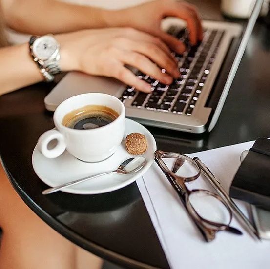 A person is typing on a laptop next to a cup of coffee on a saucer.