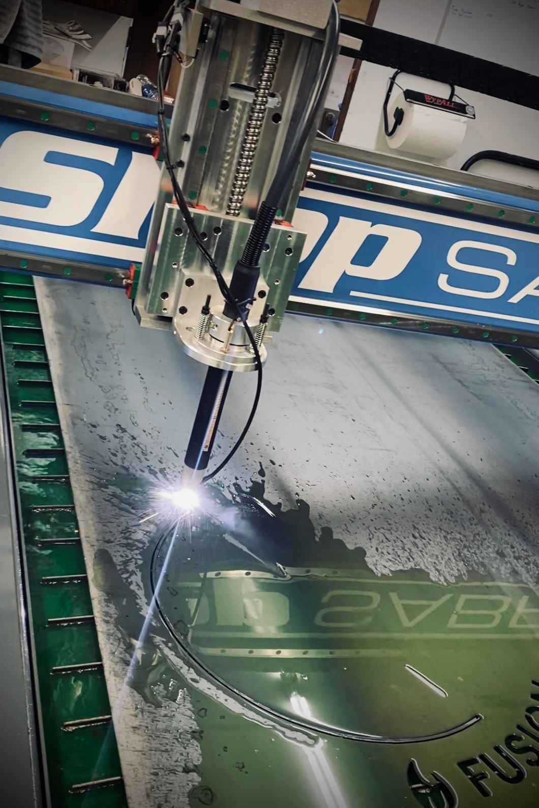 A metal sheet being cut by a machine with a bright arc, water spraying, and a blue and white Snap-Saw sign.