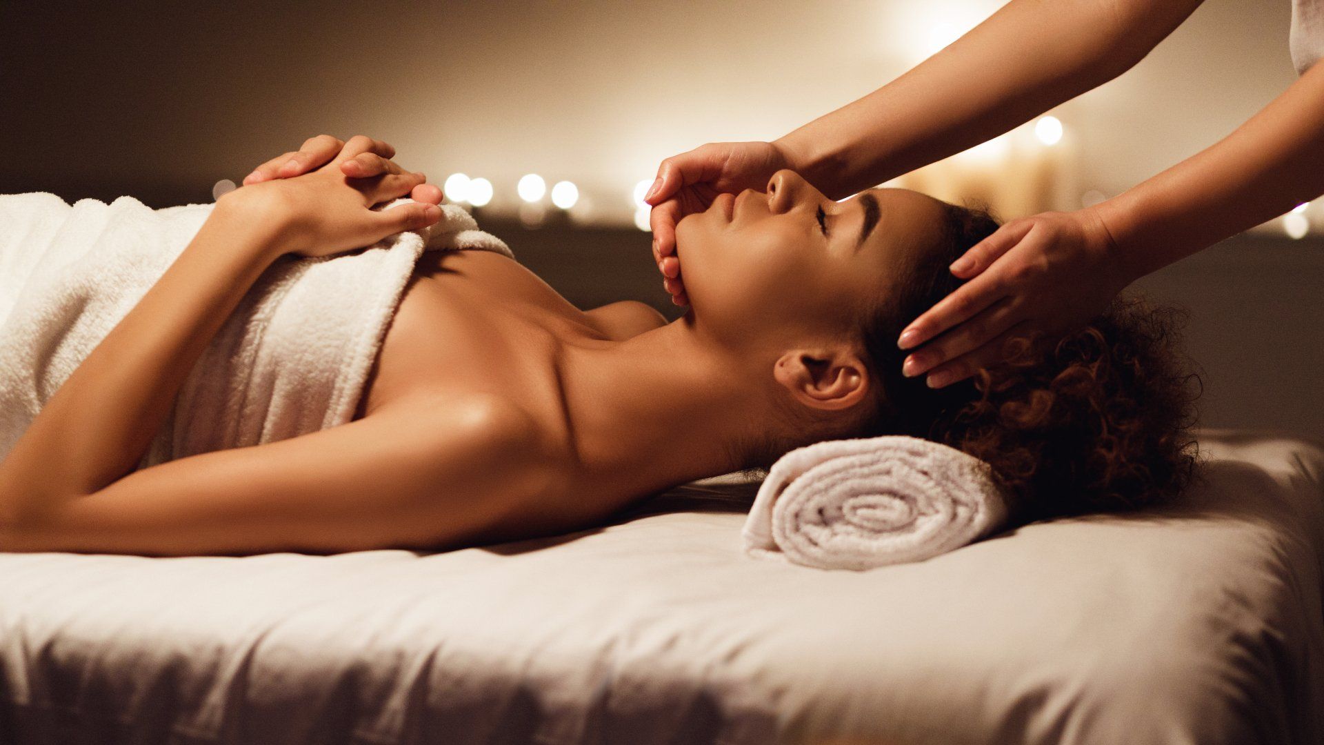 young woman laying her head on a rolled up towel on a relaxing massage bed during a massage therapy session where a massage therapist is massaging her head.