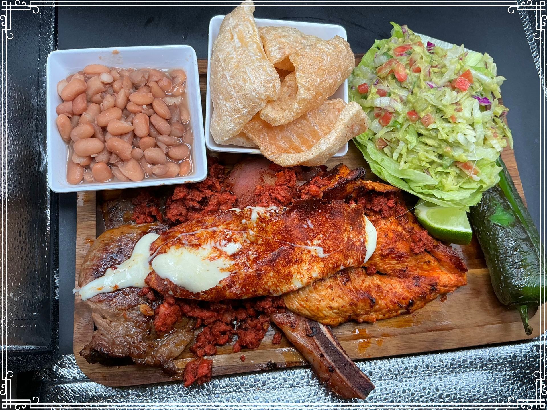 Wooden platter with cooked meats, beans, chicharrones, salad, and lime, with a dark background.