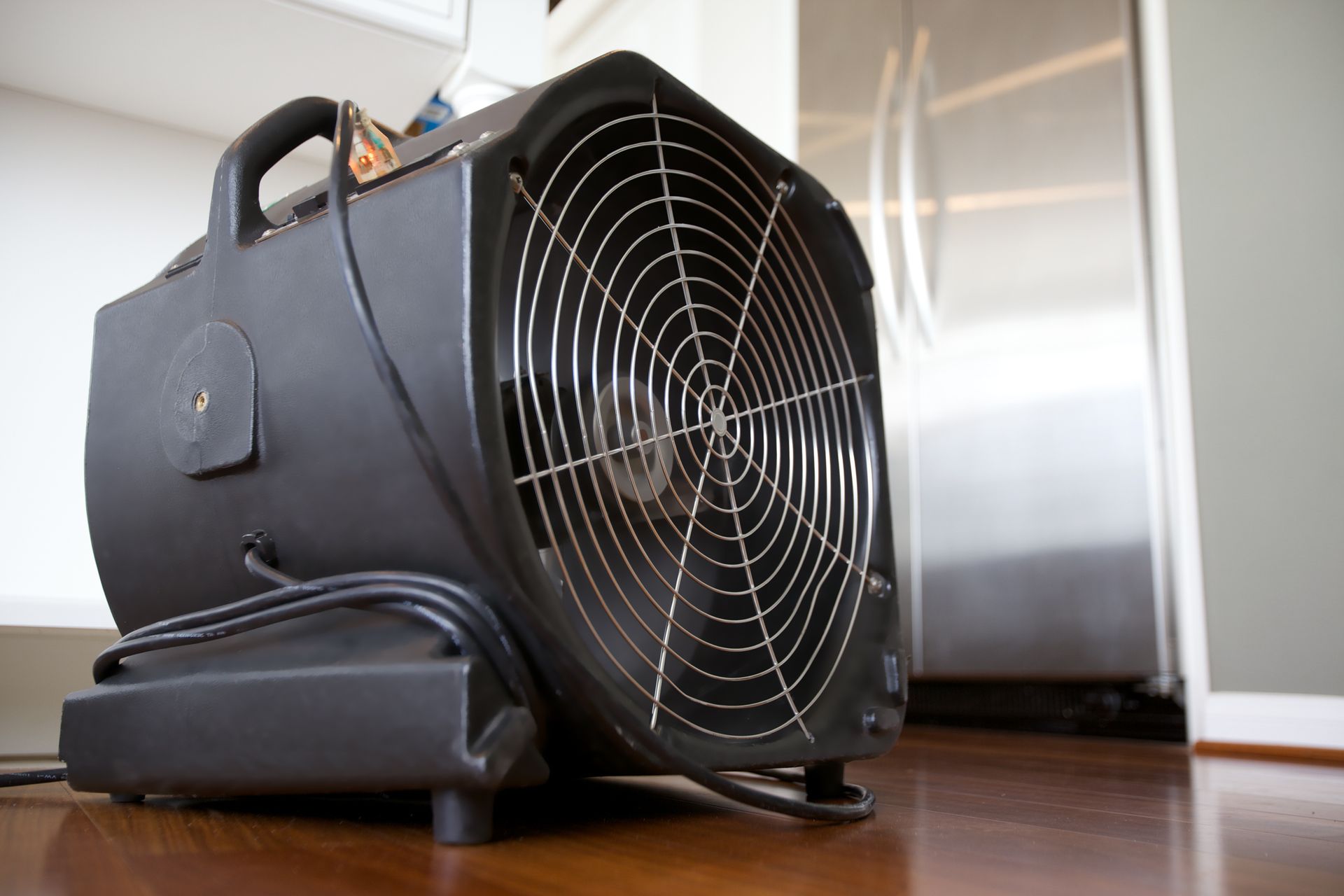 A black fan is sitting on a wooden floor in a kitchen.