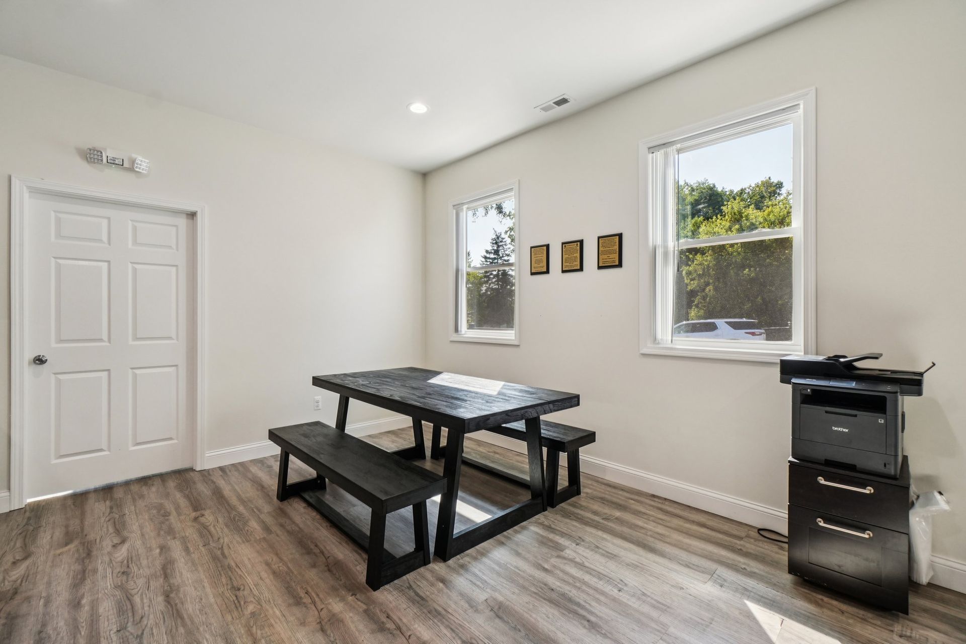 A room with a black picnic table, two windows, a printer, and a white door.