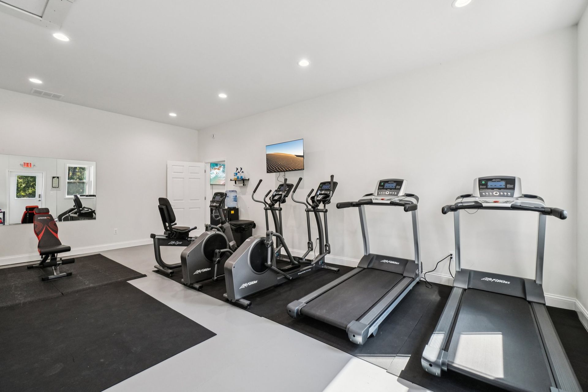 Gym with treadmills, elliptical machines, a recumbent bike, and weights on a black and grey floor.
