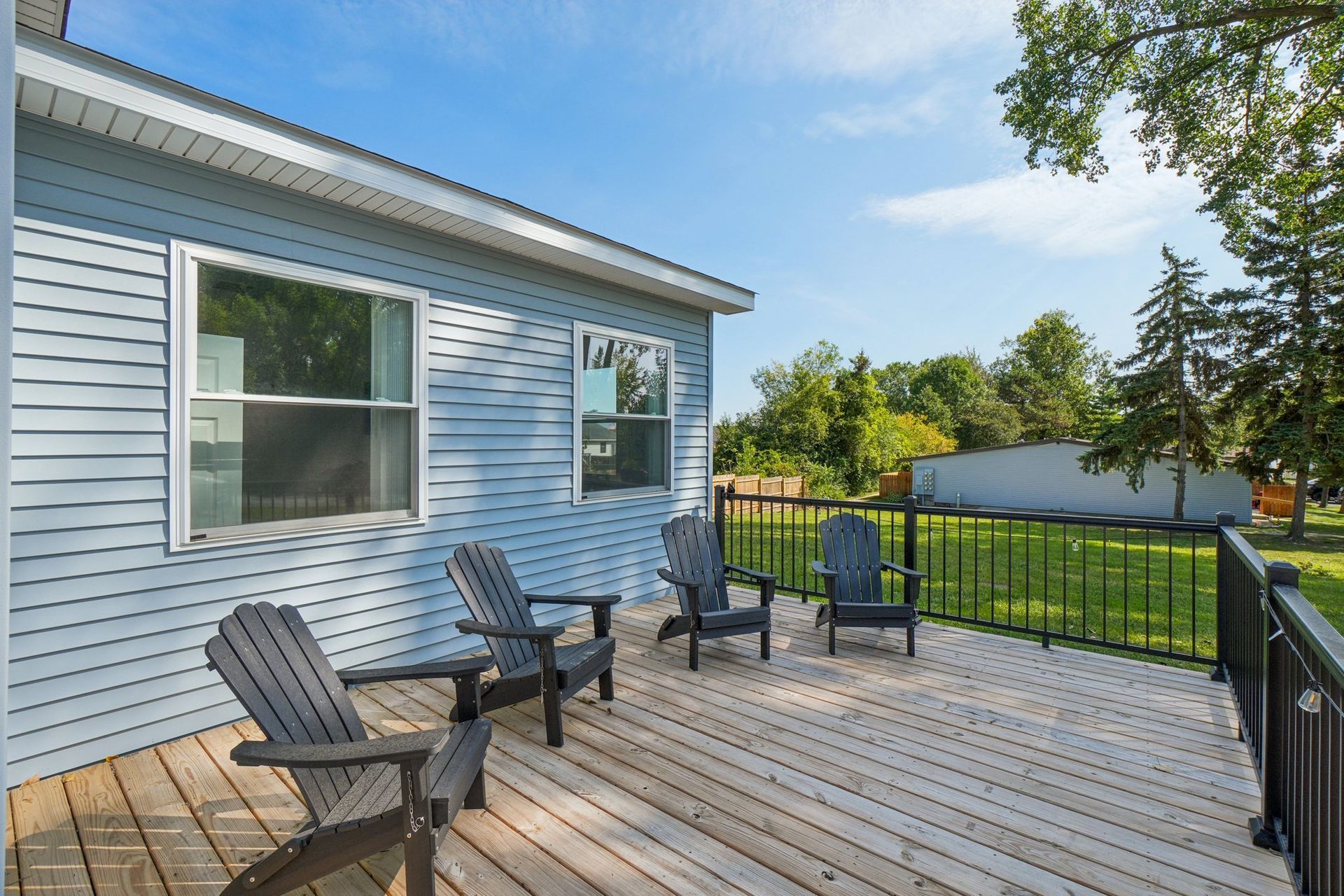Wooden deck with Adirondack chairs, next to a light blue house, and a green yard under a blue sky.