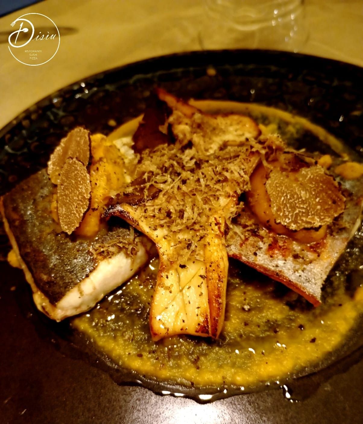 Piatto di pesce scottato in padella, guarnito con scaglie e fette di tartufo, in salsa al burro. Piatto scuro su tavolo di legno.