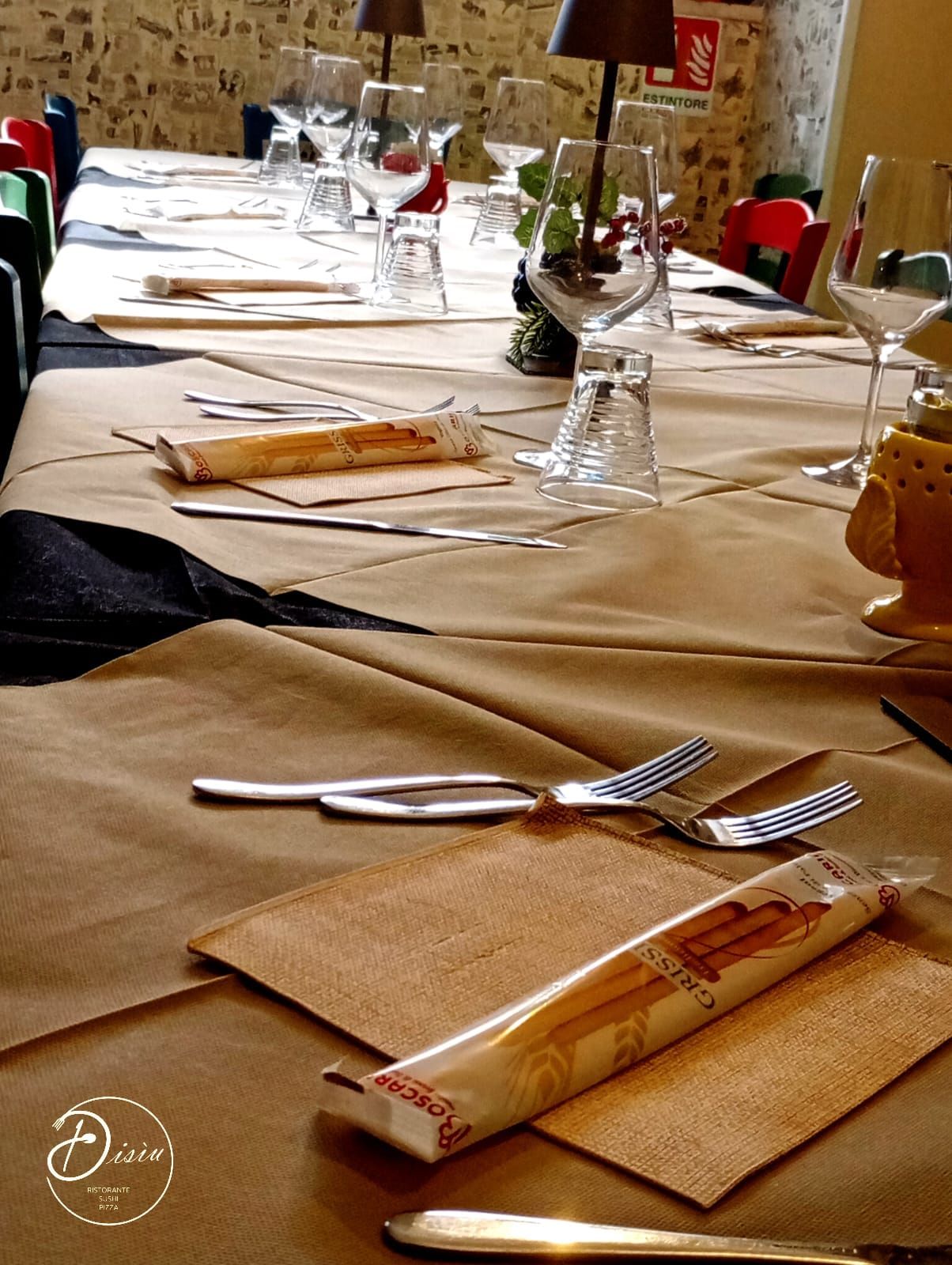 Tavolo del ristorante apparecchiato con bicchieri, posate, grissini e tovagliette di carta marrone.