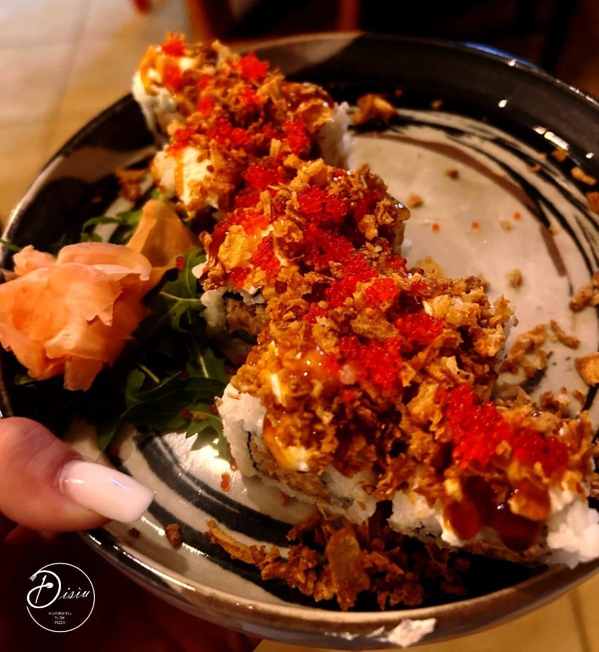 Rotolo di sushi guarnito con uova di pesce rosse e guarnizione croccante, su un piatto decorativo tenuto da una persona.