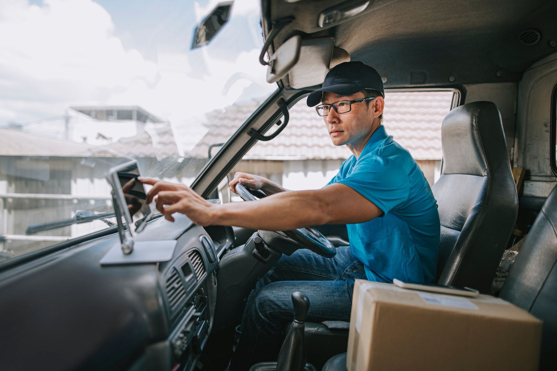 Man in a truck's driver seat, wearing a blue shirt and cap, looking at a tablet; a box sits on the passenger seat.