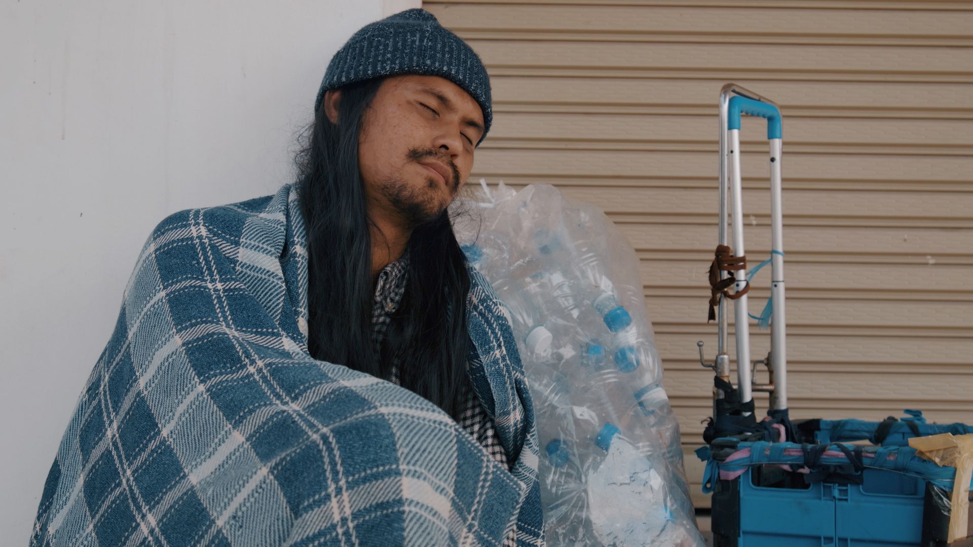 Person sleeping on a park bench, head on pillow, with belongings and a bag nearby.