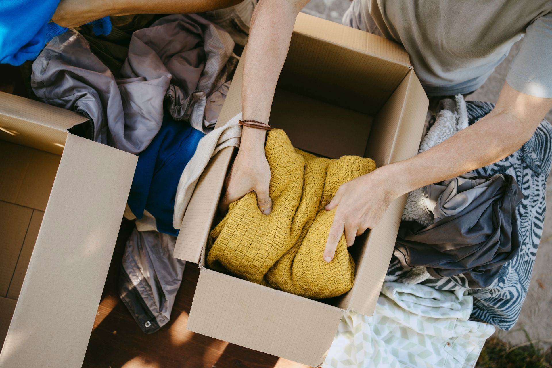 Person packing clothes into a cardboard box outdoors. Yellow sweater is being folded.