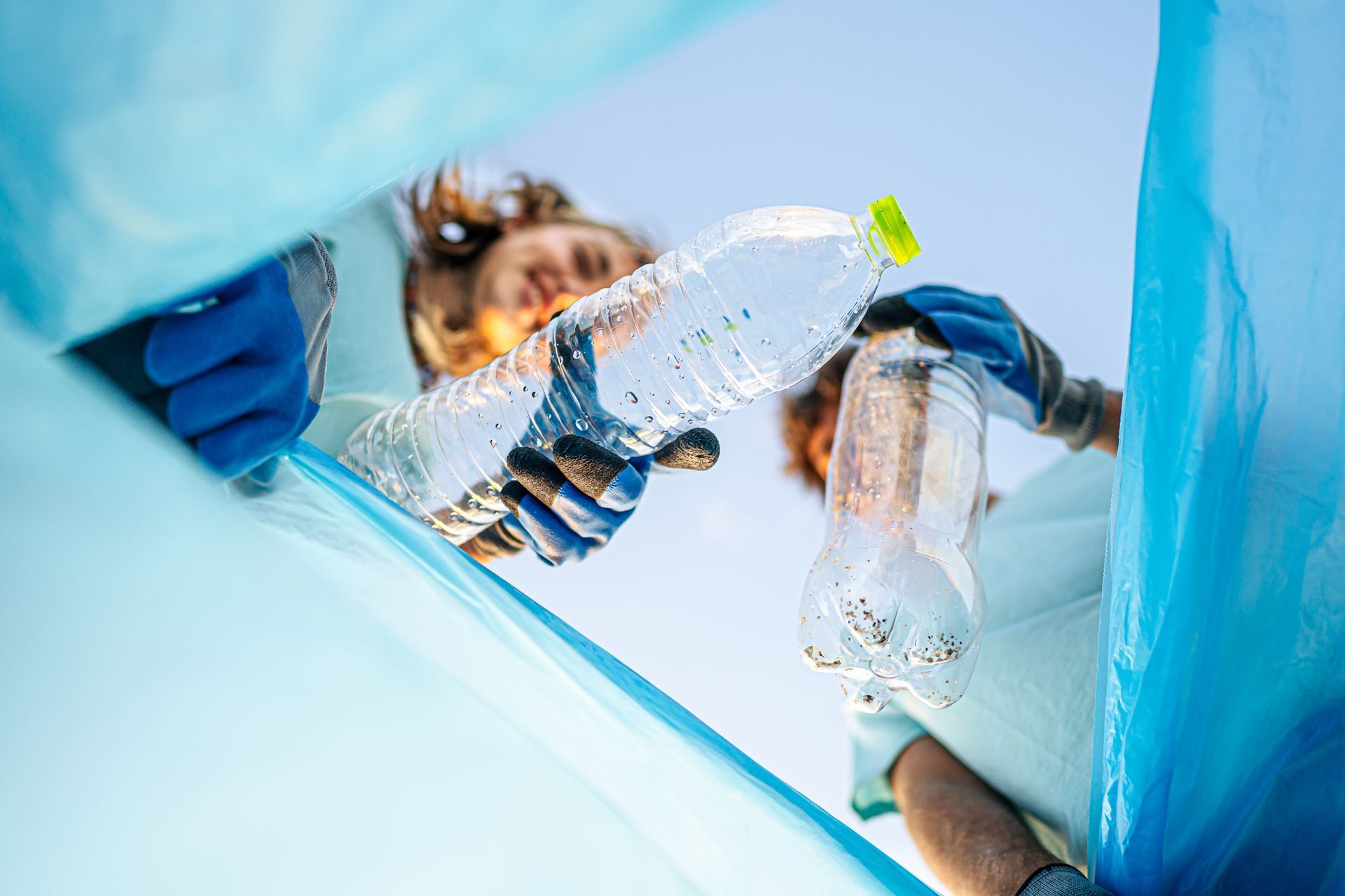 People in gloves holding plastic bottles, placing them into a blue trash bag, outdoors.