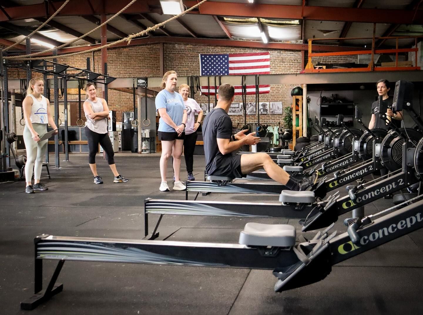A group of people are standing around rowing machines in a gym.