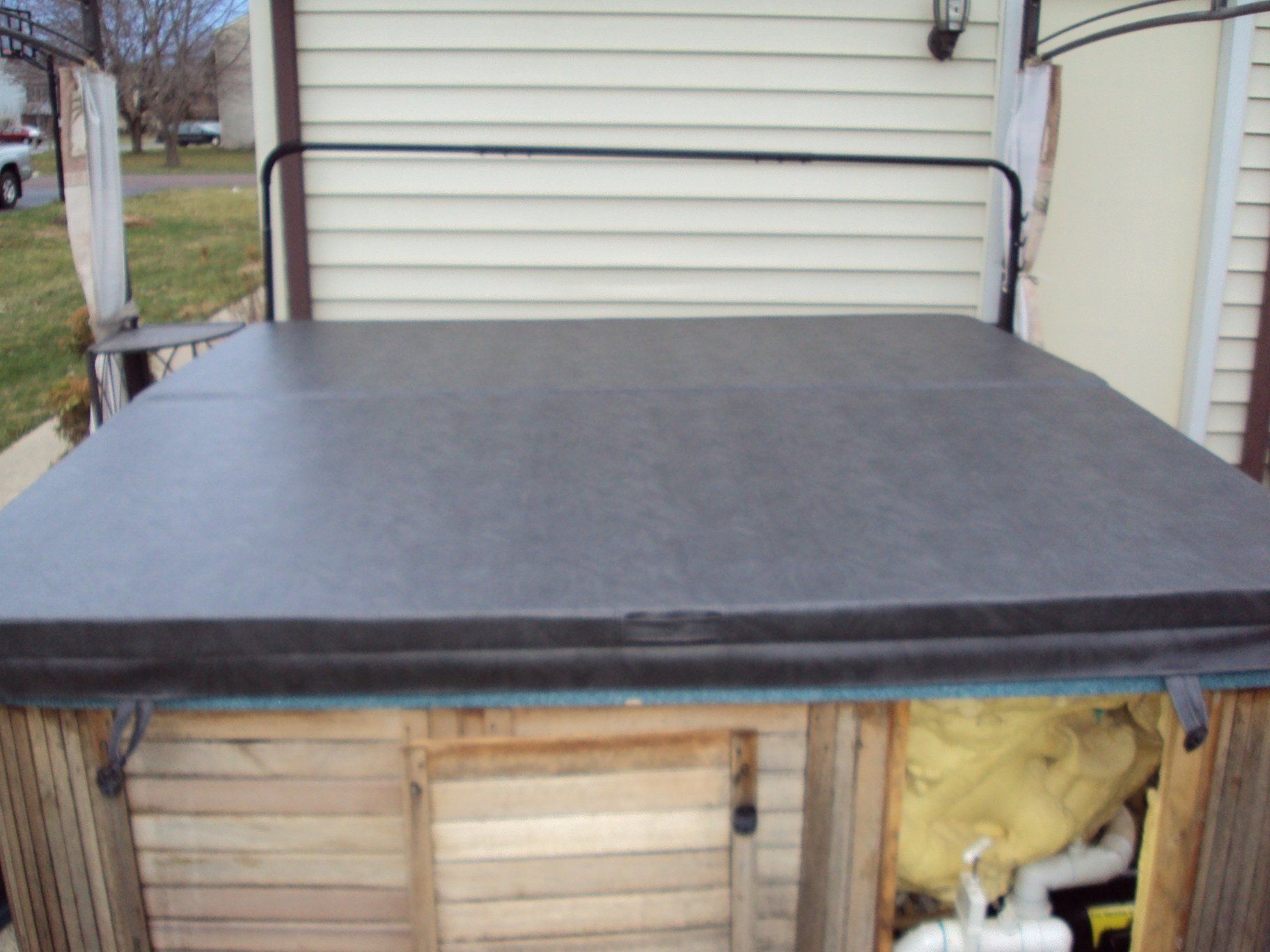 Hot tub with closed black cover, wooden frame, under a cream siding.
