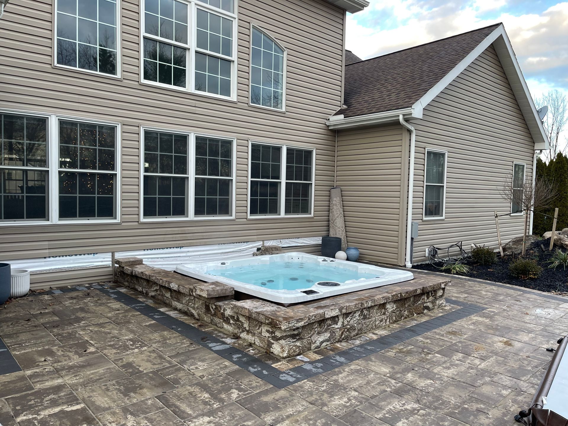 Hot tub built into a stone patio beside a two-story beige house with multiple windows.