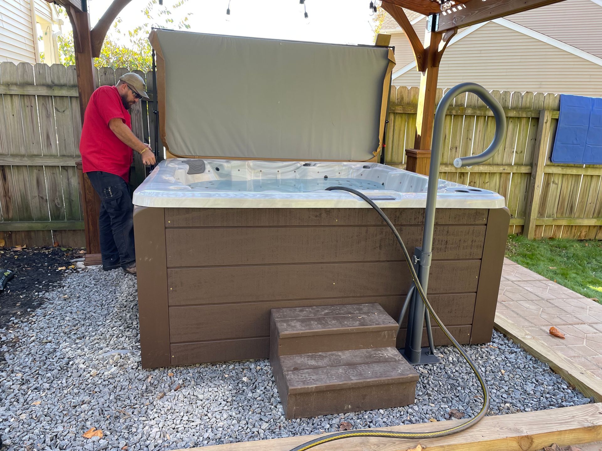 Man adjusting hot tub cover in a backyard. Brown hot tub, steps, and gazebo.