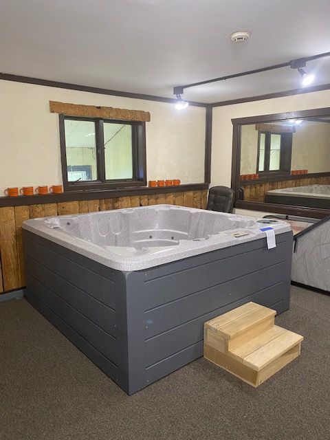 Gray hot tub with wooden steps inside a room with dark wood accents and a window.
