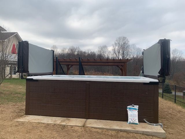 Brown hot tub with open grey covers, set outdoors on concrete pads, with a cloudy sky.