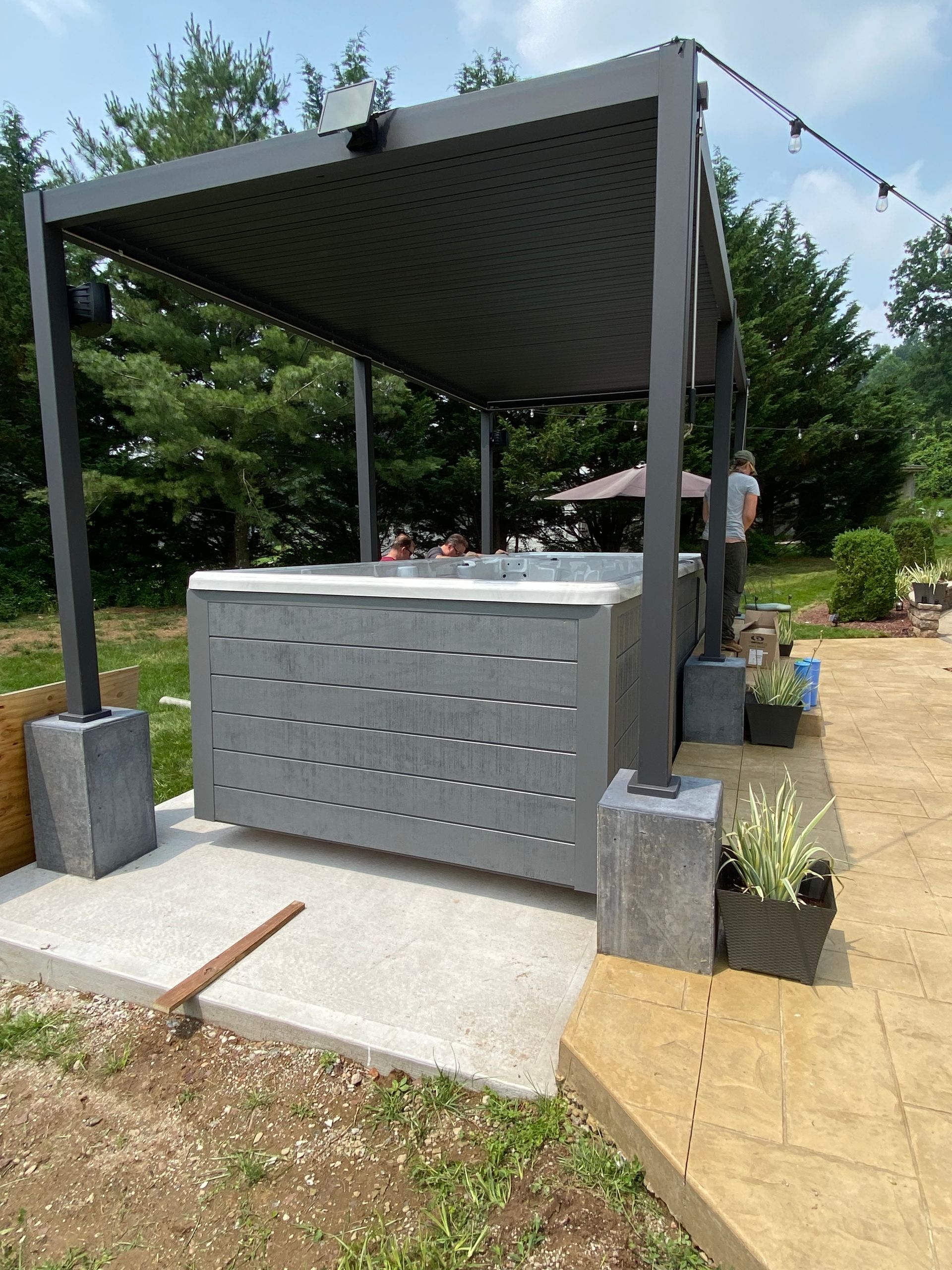 A gray hot tub under a black pergola on a concrete patio, with plants and string lights.