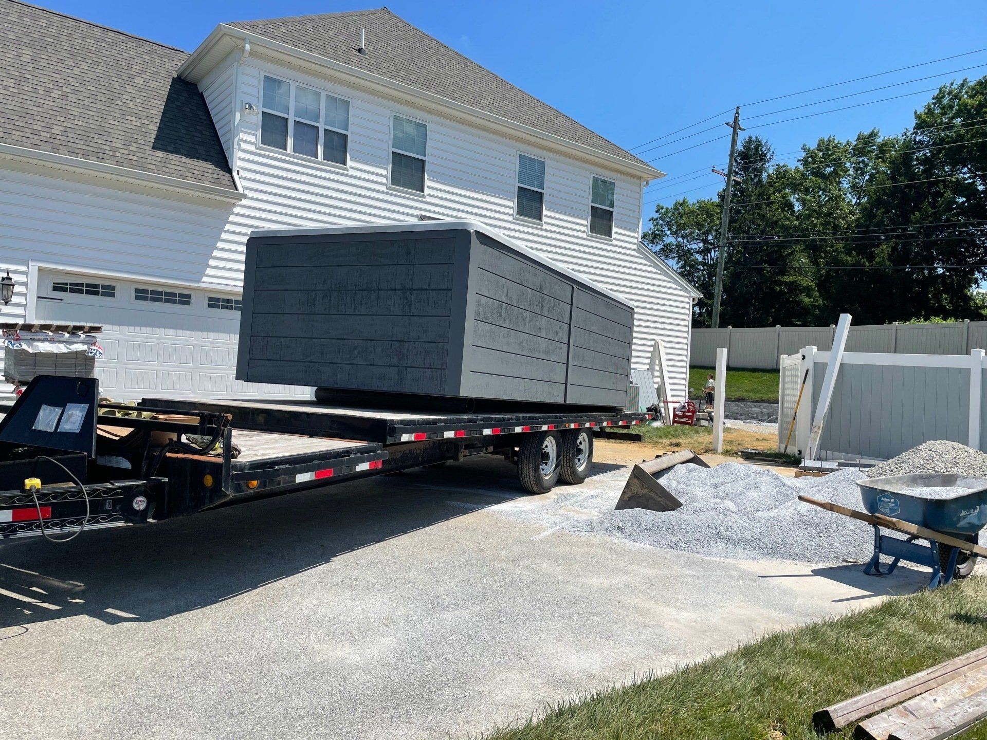 A flatbed trailer holding dark blocks, parked in front of a two-story house with construction materials nearby.
