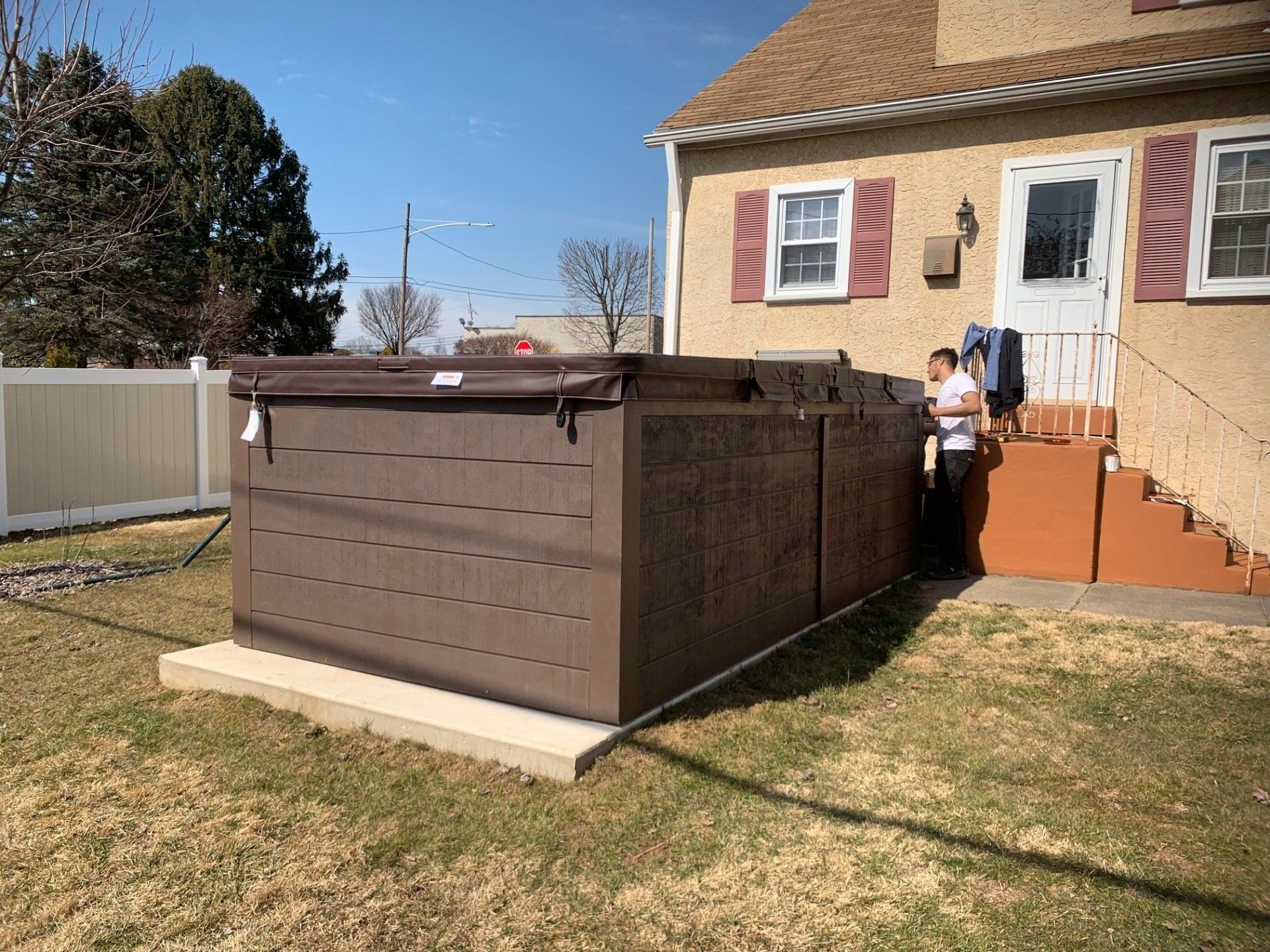 Hot tub beside a house. Brown enclosure with the cover open; person stands nearby.