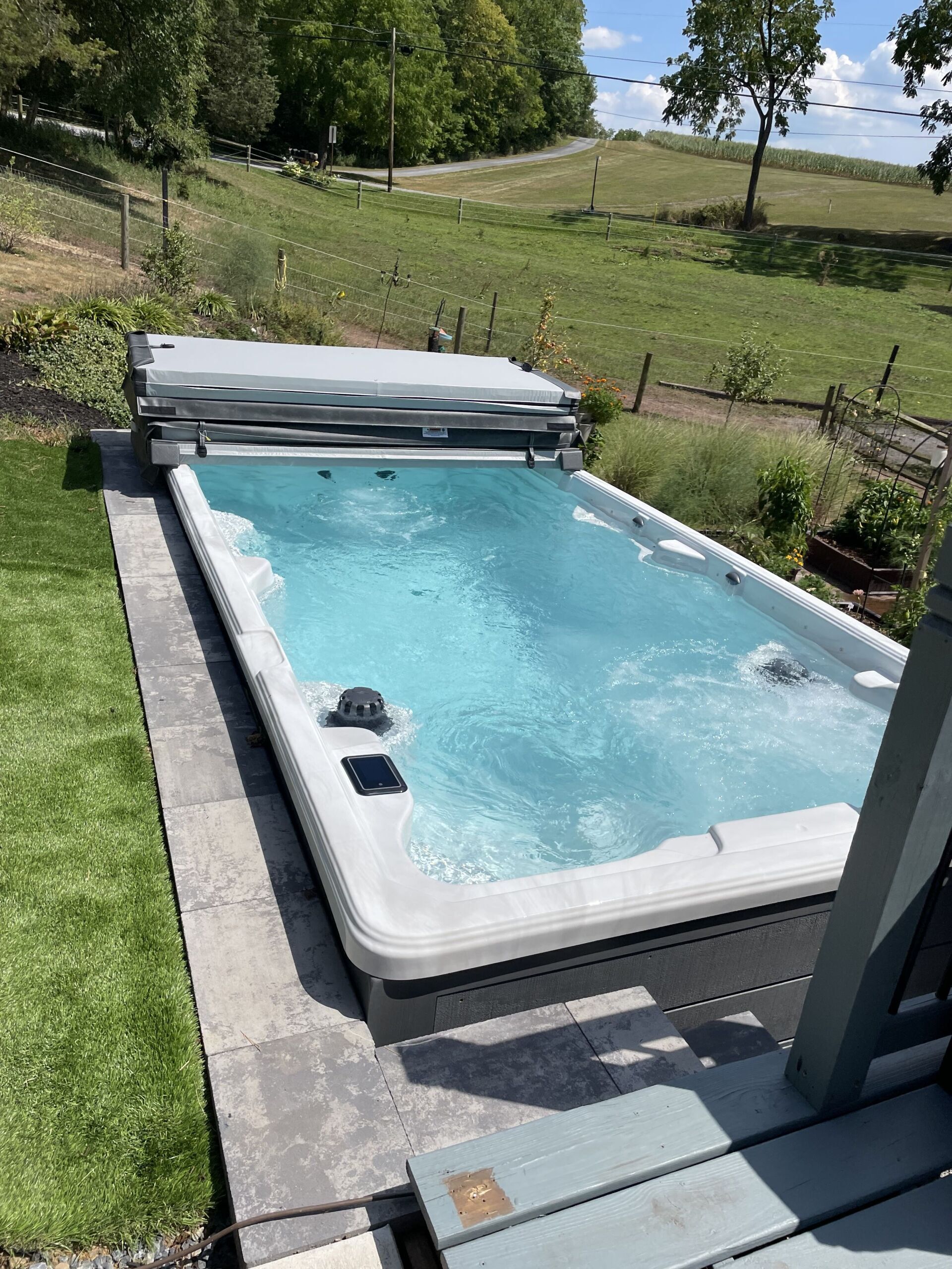 A long, rectangular white swim spa filled with blue water, set outdoors near green grass and a lake.
