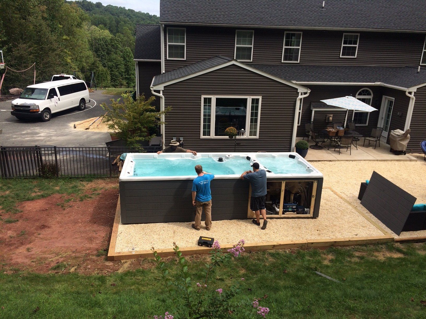 Two men near a dark gray hot tub in a backyard. White van, house, and patio visible.