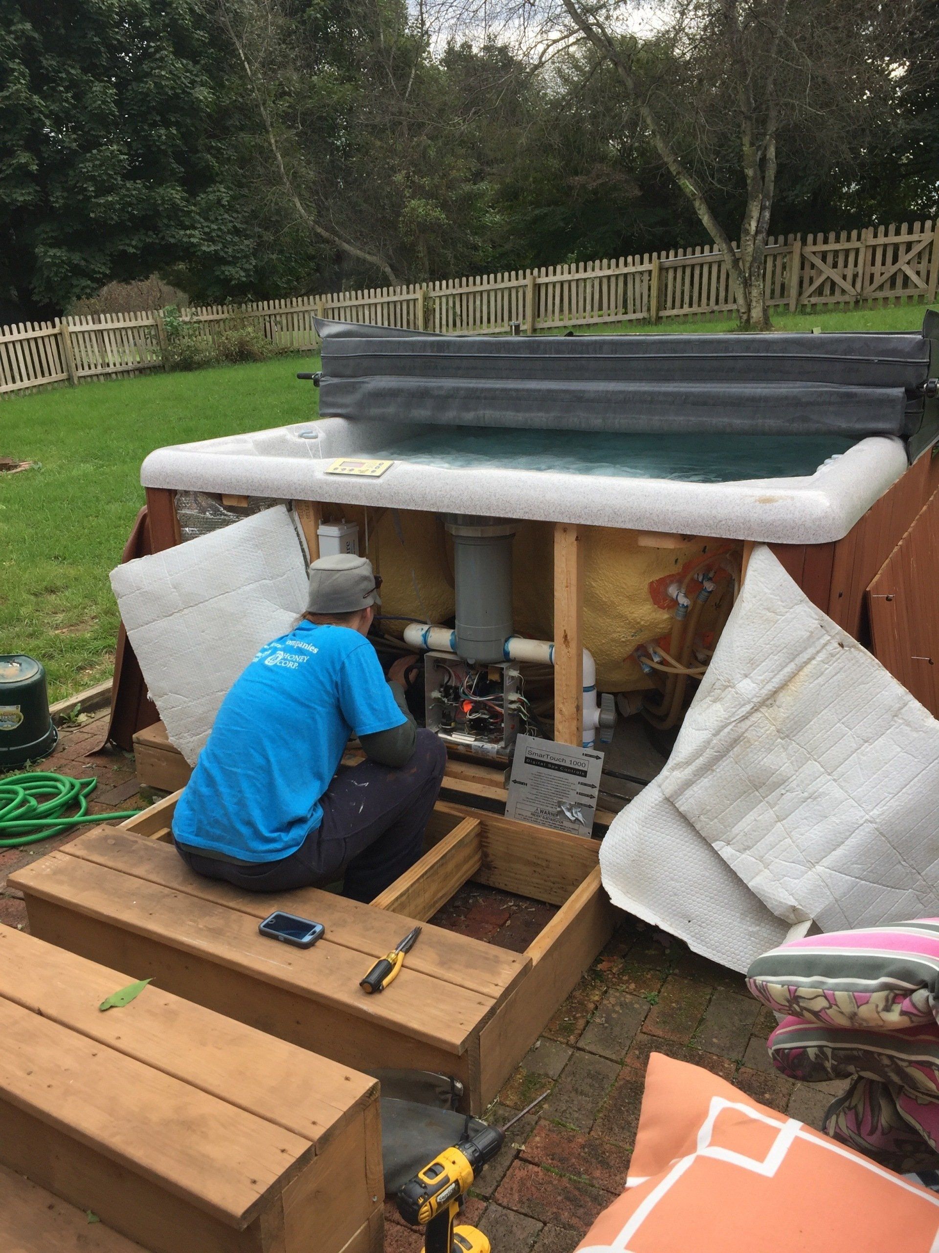 A person in a blue shirt repairs a hot tub. Exterior setting with a wooden deck, backyard, and a fence.