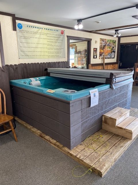 Hot tub display in showroom, brown exterior, blue interior, wooden steps, beige rug.