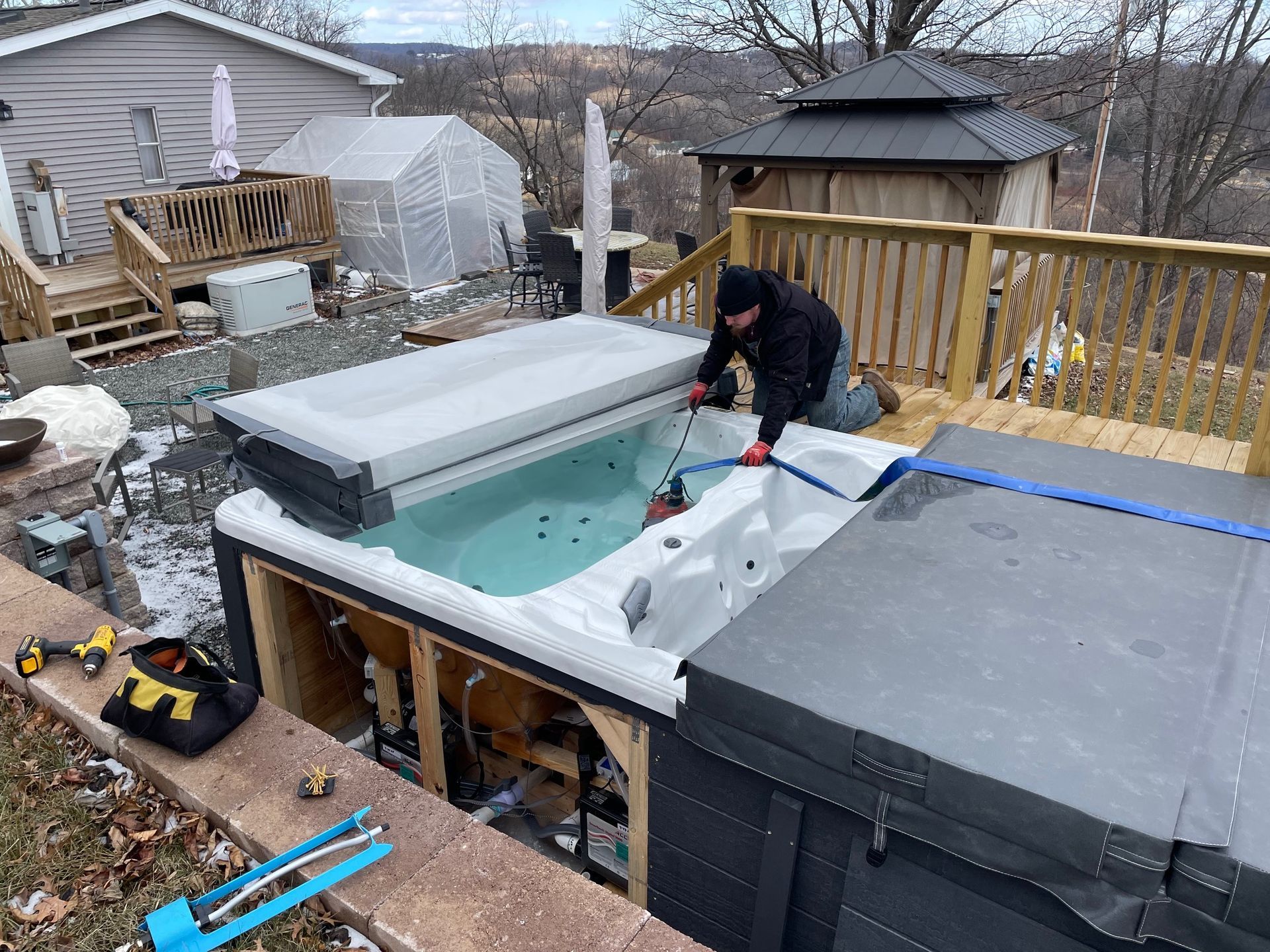 Man cleaning a hot tub on a wooden deck; a gazebo and greenhouse are in the background.