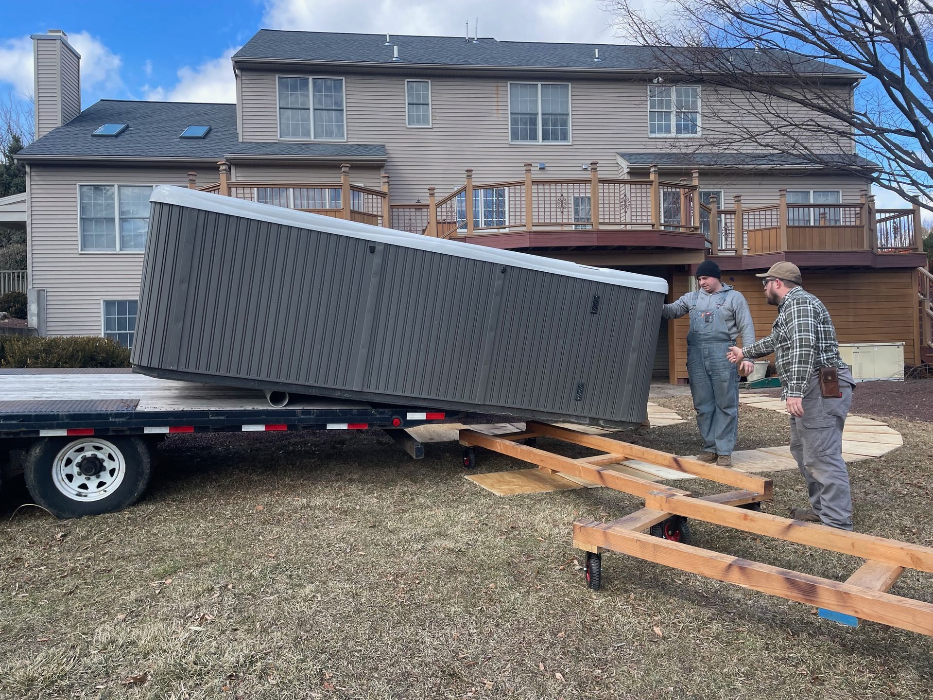 Two workers unloading a hot tub from a trailer in a backyard, house in background.