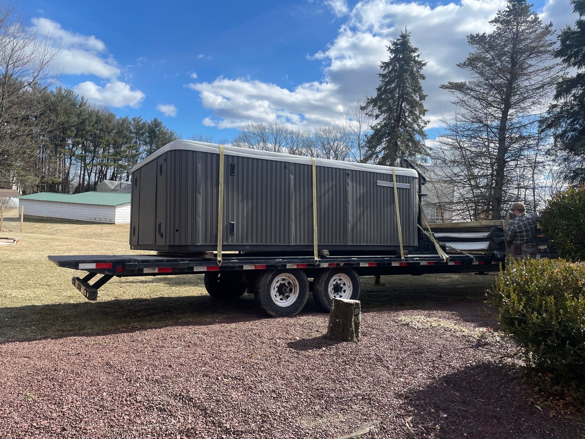 A large gray structure secured on a flatbed trailer, parked on grass. Blue sky and trees in the background.