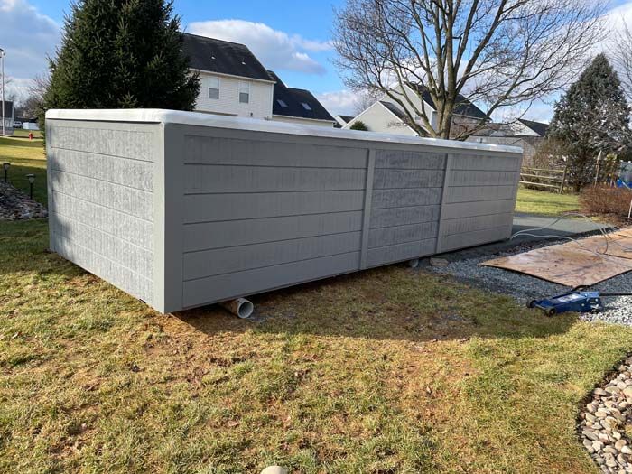 Gray rectangular storage shed in a yard, near a house, on a sunny day.