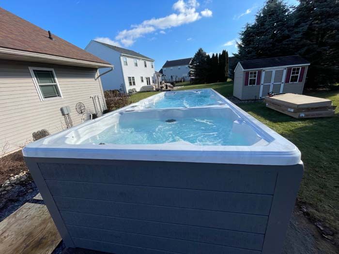Long, rectangular swim spa in backyard, blue water, gray exterior, sunny day.