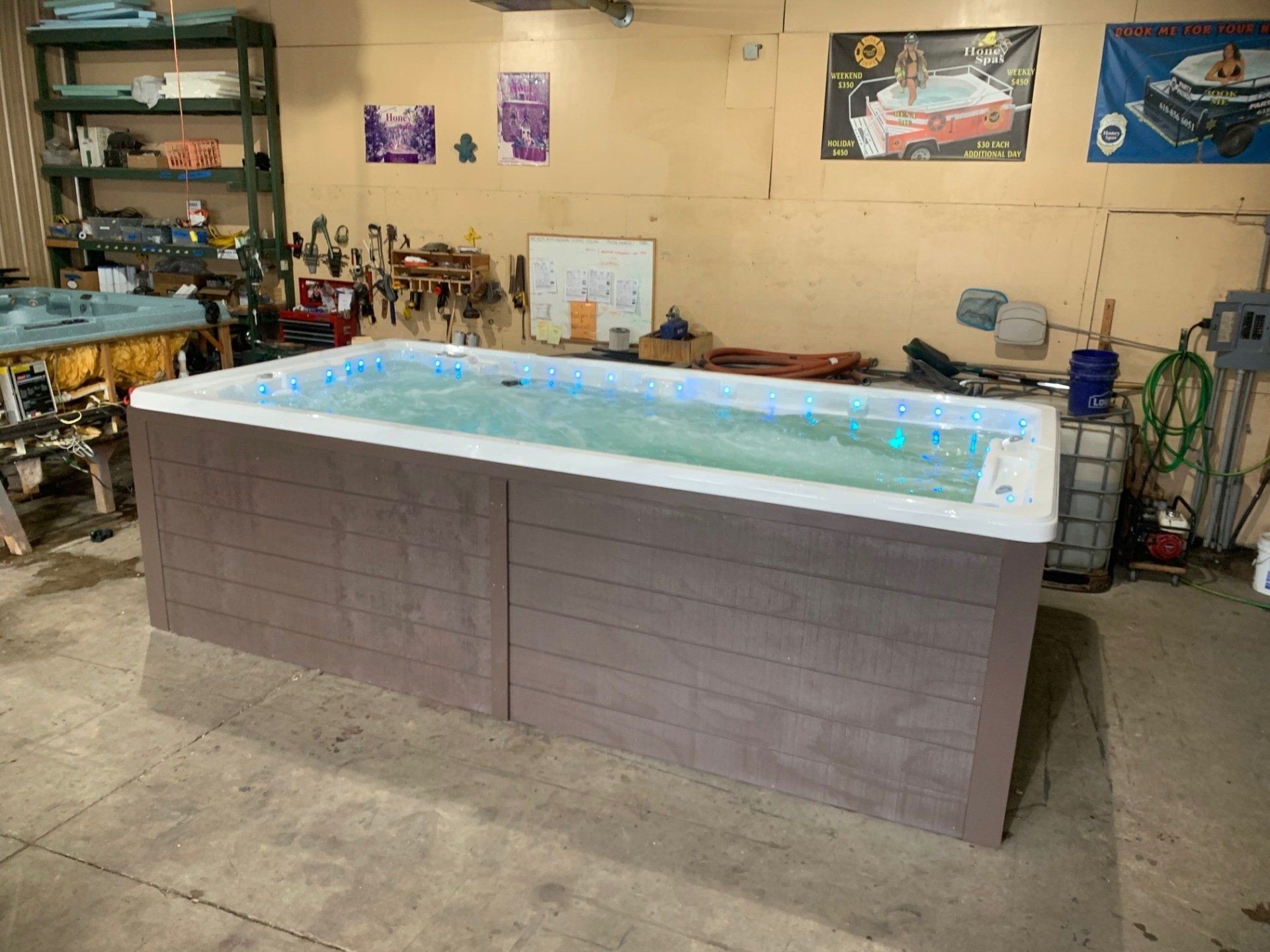 A large, rectangular swim spa indoors; wooden siding, water with lights, set in a garage with tools and equipment.