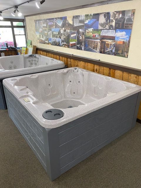 A square grey and white hot tub sits inside a showroom with a picture collage backdrop.