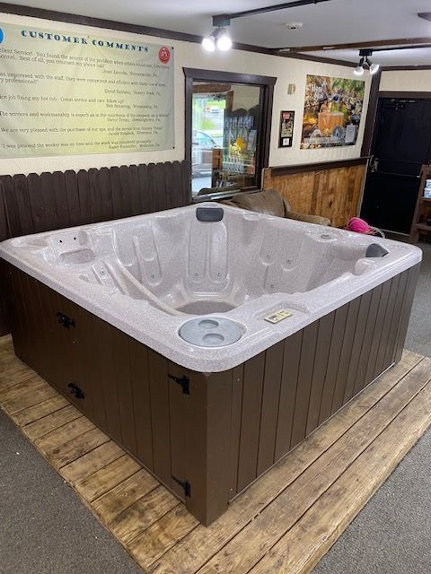 Brown hot tub in a store showroom on a wooden pallet, with wall of customer testimonials.