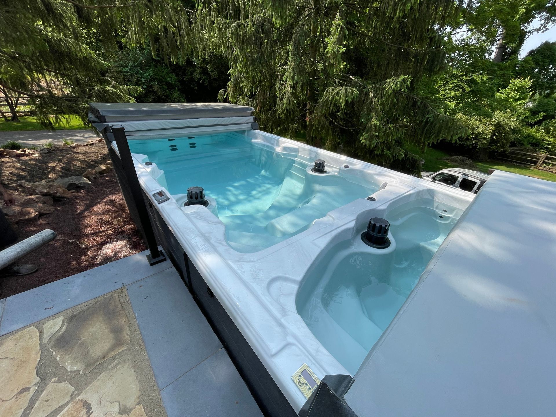 Swim spa outdoors with clear water and cover partially open, surrounded by greenery and stone patio.