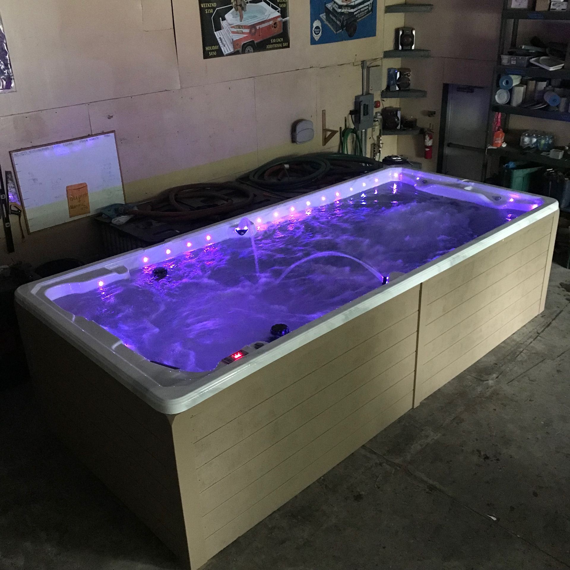 Swim spa outdoors with clear water and cover partially open, surrounded by greenery and stone patio.