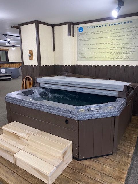 A brown hot tub with water, steps, and a partially open cover, indoors with a customer comments sign.