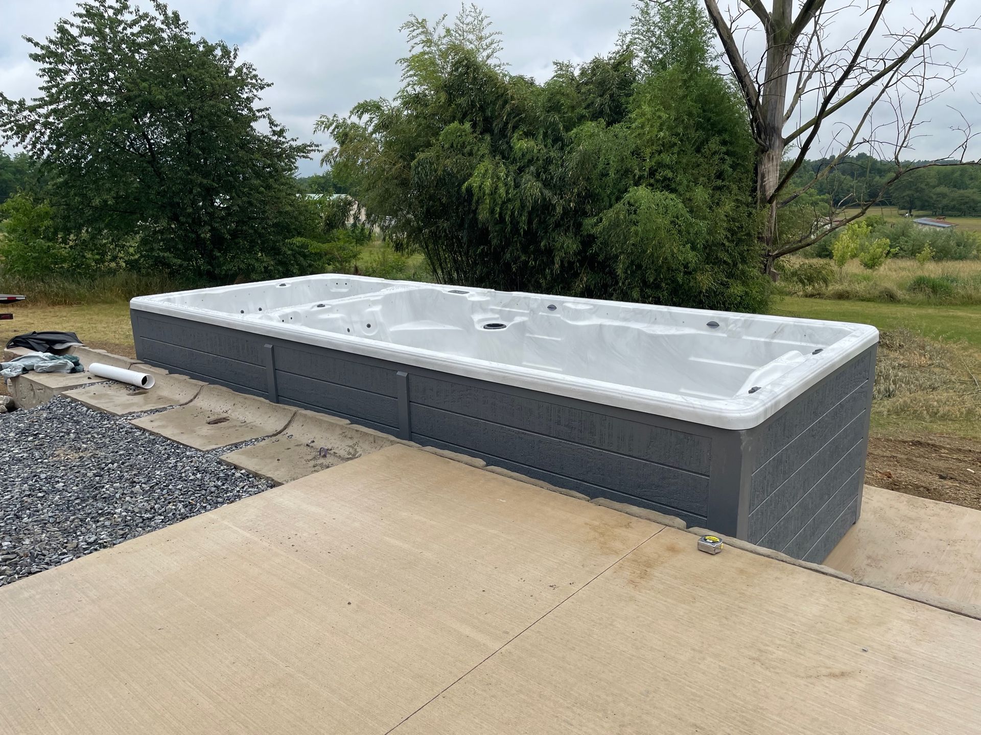 A rectangular gray swim spa on a concrete patio, surrounded by a grassy yard and trees.