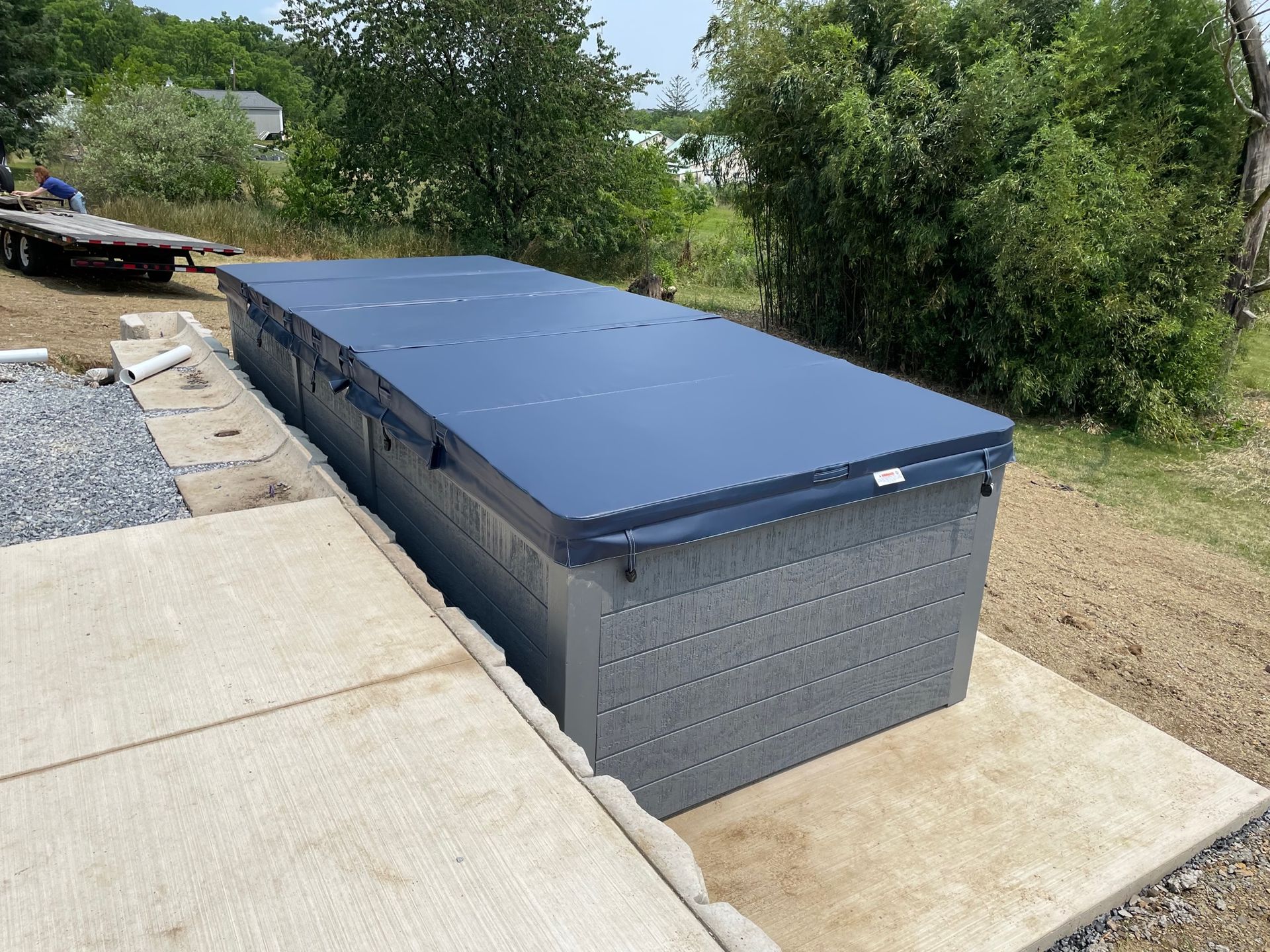 A long, rectangular hot tub with a brown cover on an asphalt lot, next to a building.