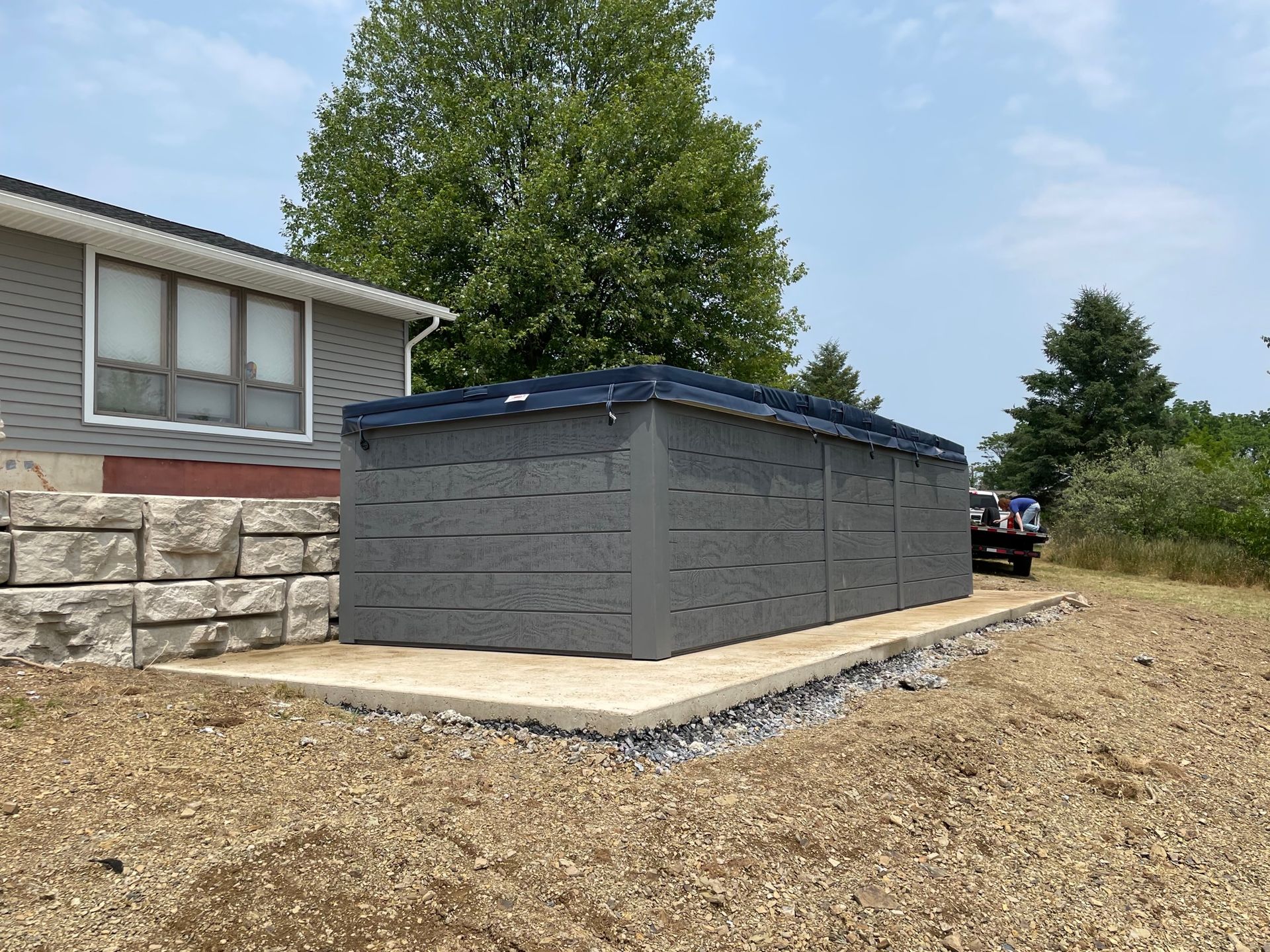 Gray shed on a concrete pad next to a house. Gravel and retaining wall are in the foreground.