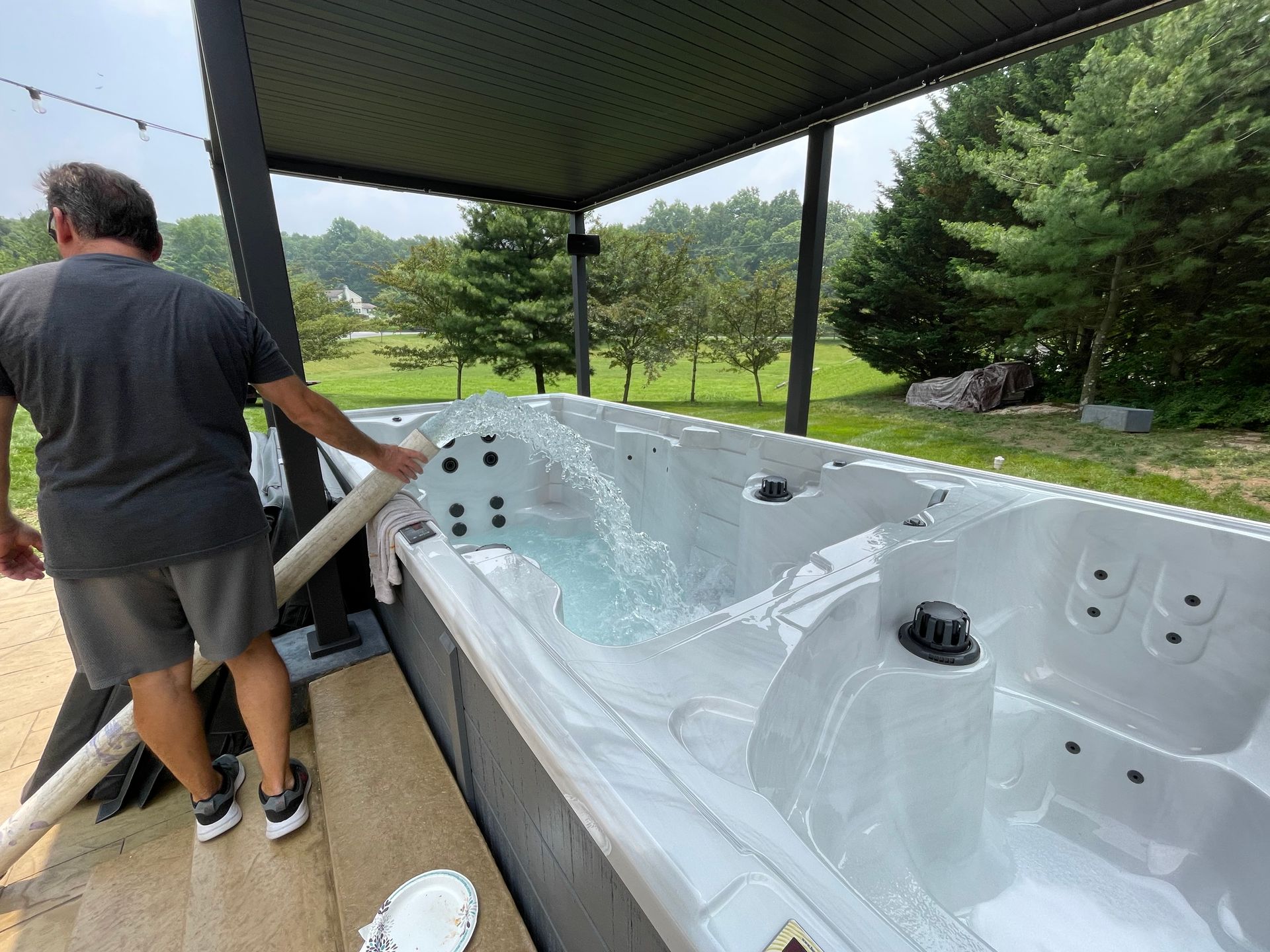 Man filling a swim spa with water outdoors. Spa is under a pergola, green trees in background.