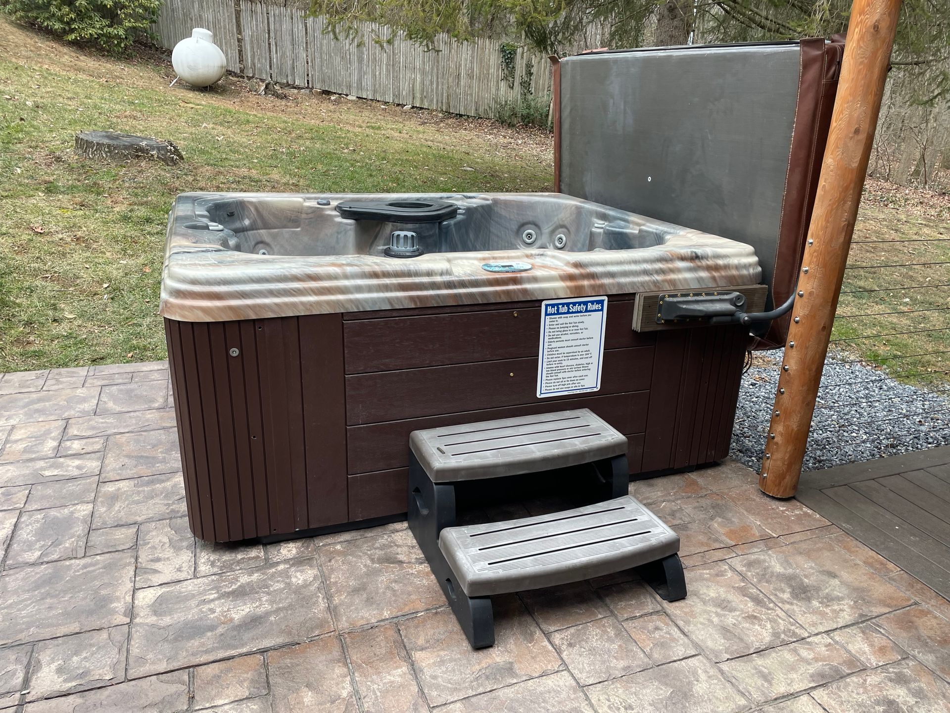 A brown hot tub with the cover open sits on a stone patio with gray steps in front.