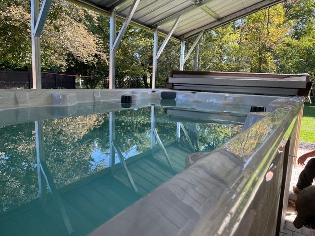 Long, rectangular swim spa under a metal shelter, filled with water; surrounded by grass and trees.
