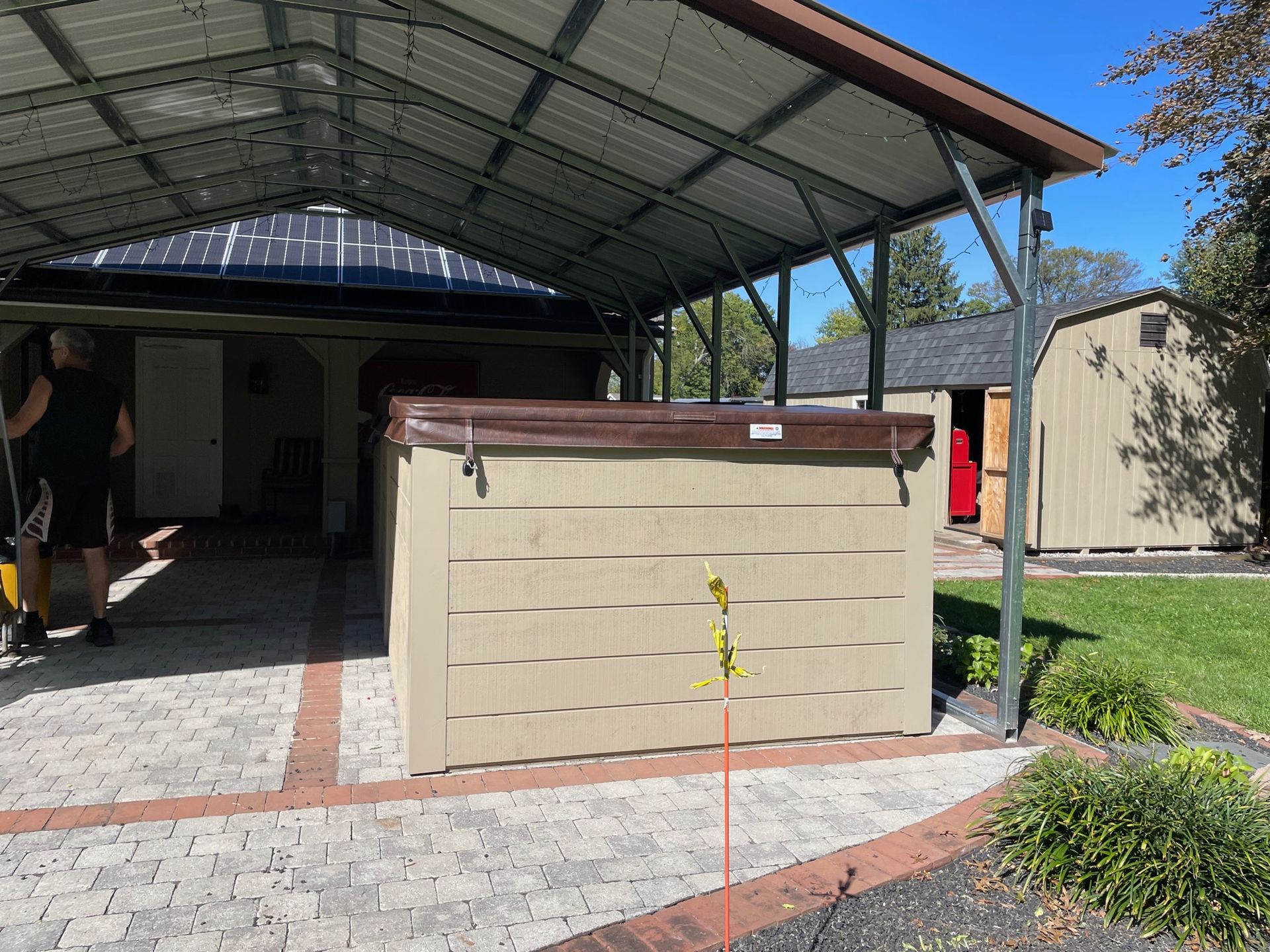 Hot tub under a metal roof, set on brick patio near a shed, person stands nearby.