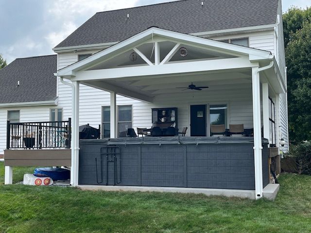 Covered outdoor deck with spa, attached to a two-story white house. Gray siding and a green lawn.