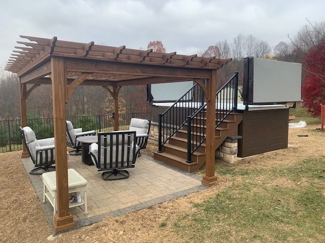 A pergola with seating next to a hot tub with steps and black railing, on a patio with a grassy background.