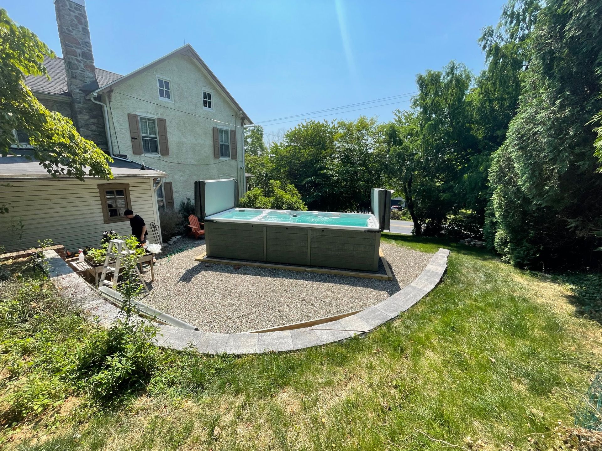 Backyard scene: rectangle pool on gravel bed, near a house with trees and blue sky.