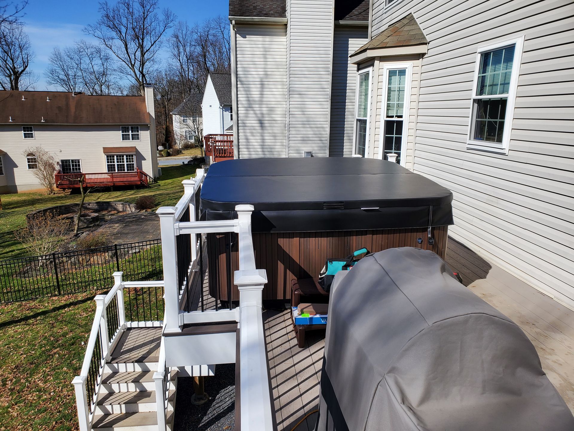 Hot tub on a wooden deck next to a house, trees in the background, and a clear sky.